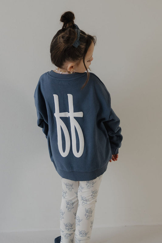 A young girl with dark hair in a bun wears the forever french baby Child Sweatshirt | ff Blue, featuring large white abstract letters on the back, paired with white leggings with a blue floral pattern as she stands facing away from the camera.