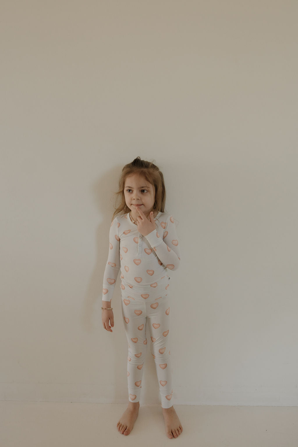 A young girl stands barefoot by a light wall in forever french baby’s Bamboo Two Piece Pajamas | Love Notes - Grey, looking to the side with one hand near her mouth.
