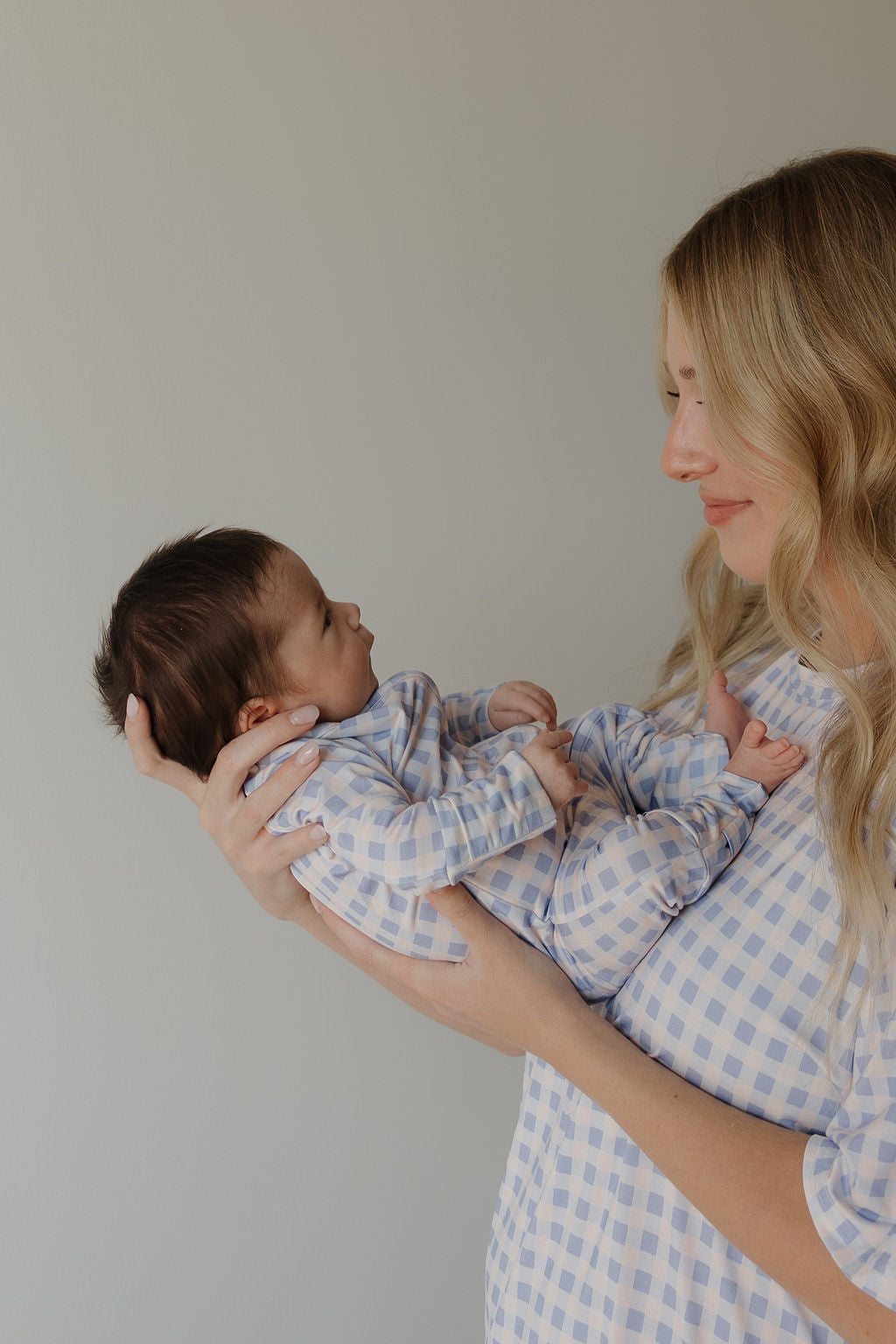 A woman with long blonde hair holds a newborn baby, both dressed in matching blue and white plaid Bamboo Zip Pajamas by forever french baby (style: Marilyn), facing each other against a plain light background in soft, breathable outfits.