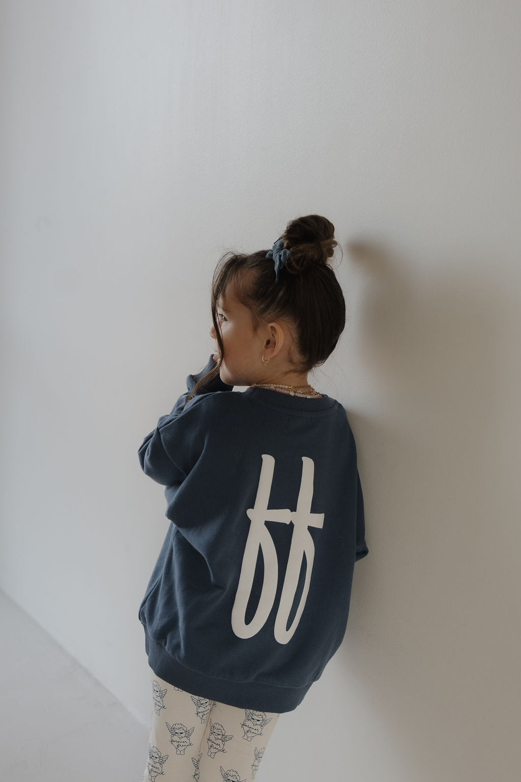 A young child with a bun stands against a white wall, wearing the forever french baby Child Sweatshirt | ff Blue with large white "ff" letters on the back and light floral print pants.