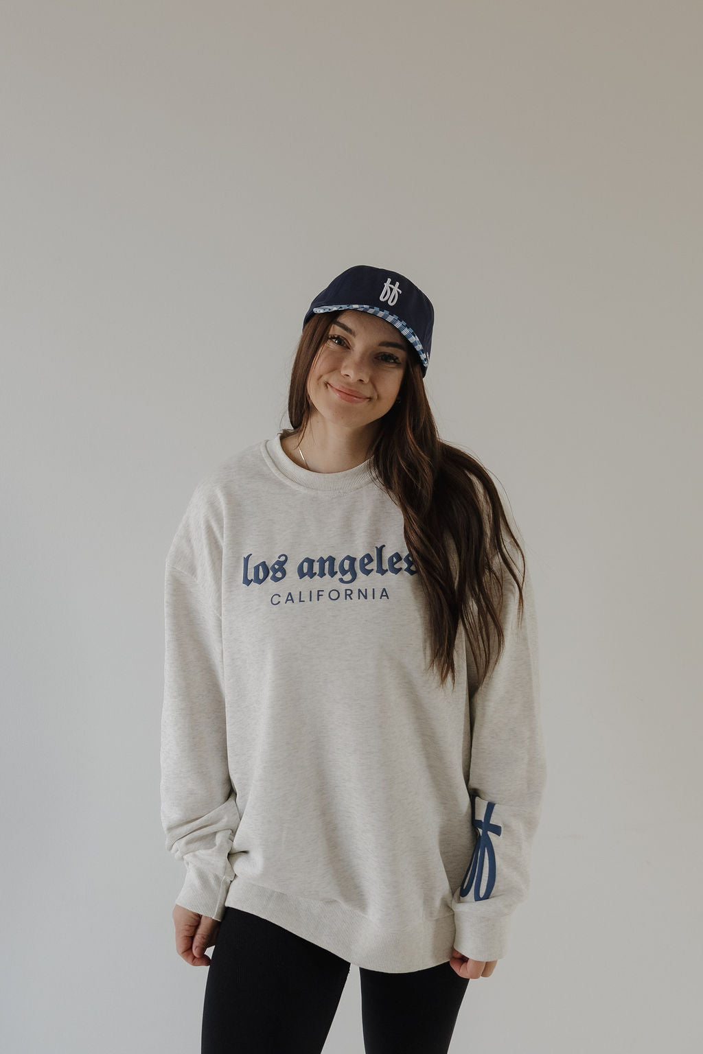 A woman smiles, standing against a light background in a relaxed-fit light gray “Adult Sweatshirt | ff Los Angeles” by forever french baby, paired with a navy blue baseball cap and her hands by her sides.