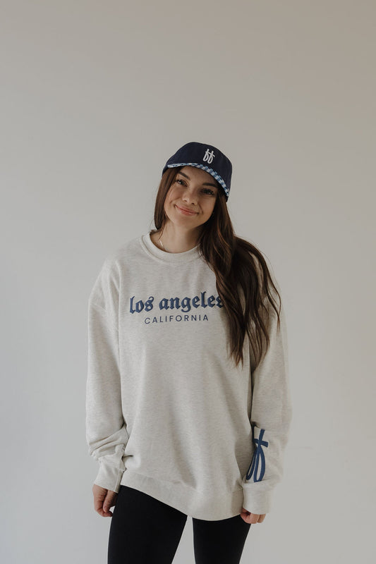 A woman smiles, standing against a light background in a relaxed-fit light gray “Adult Sweatshirt | ff Los Angeles” by forever french baby, paired with a navy blue baseball cap and her hands by her sides.