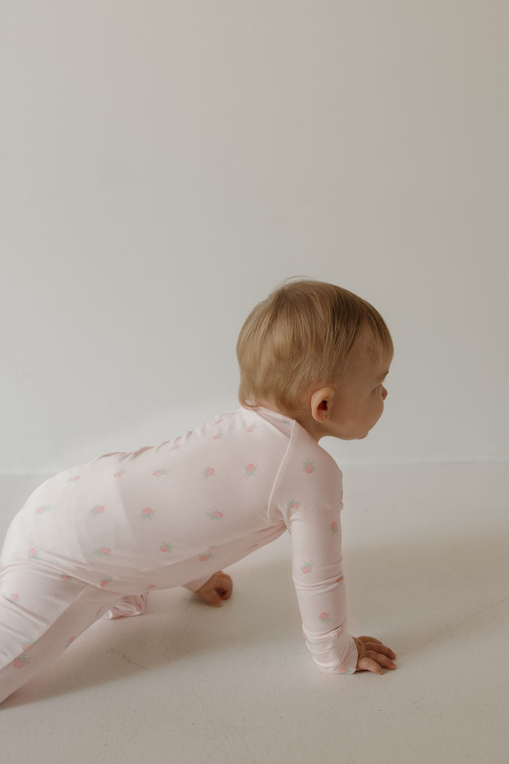 A baby wearing the forever french baby Bamboo Zip Pajamas | Rosie, featuring a light pink hue with delicate floral prints, crawls on a light-colored floor against a plain white wall, highlighting the comfort of hypoallergenic bamboo baby clothing.