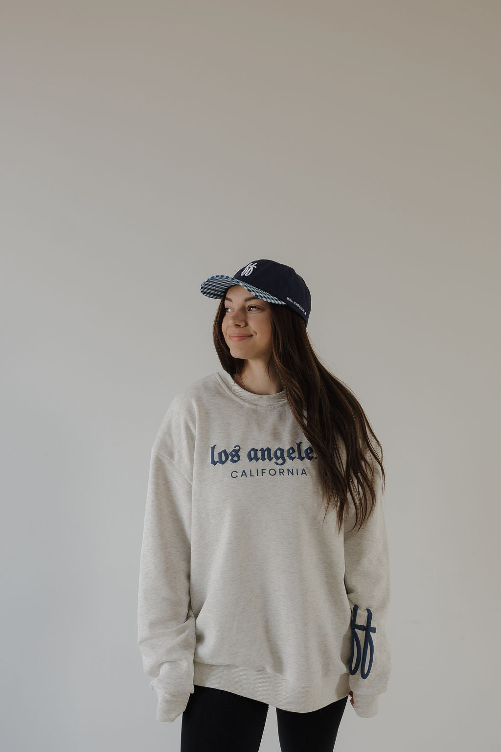 A woman with long brown hair wears a forever french baby Adult Sweatshirt | ff Los Angeles in relaxed fit light gray, along with a navy baseball cap, standing against a plain light background and smiling slightly.