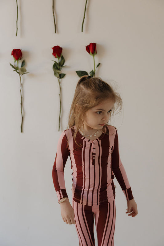A young girl models the forever french baby Bamboo Two Piece Pajamas | Sweetheart Stripes, standing by a white wall with four vertically spaced red roses. She glances to the side, showcasing these stylish bamboo pajamas for kids.