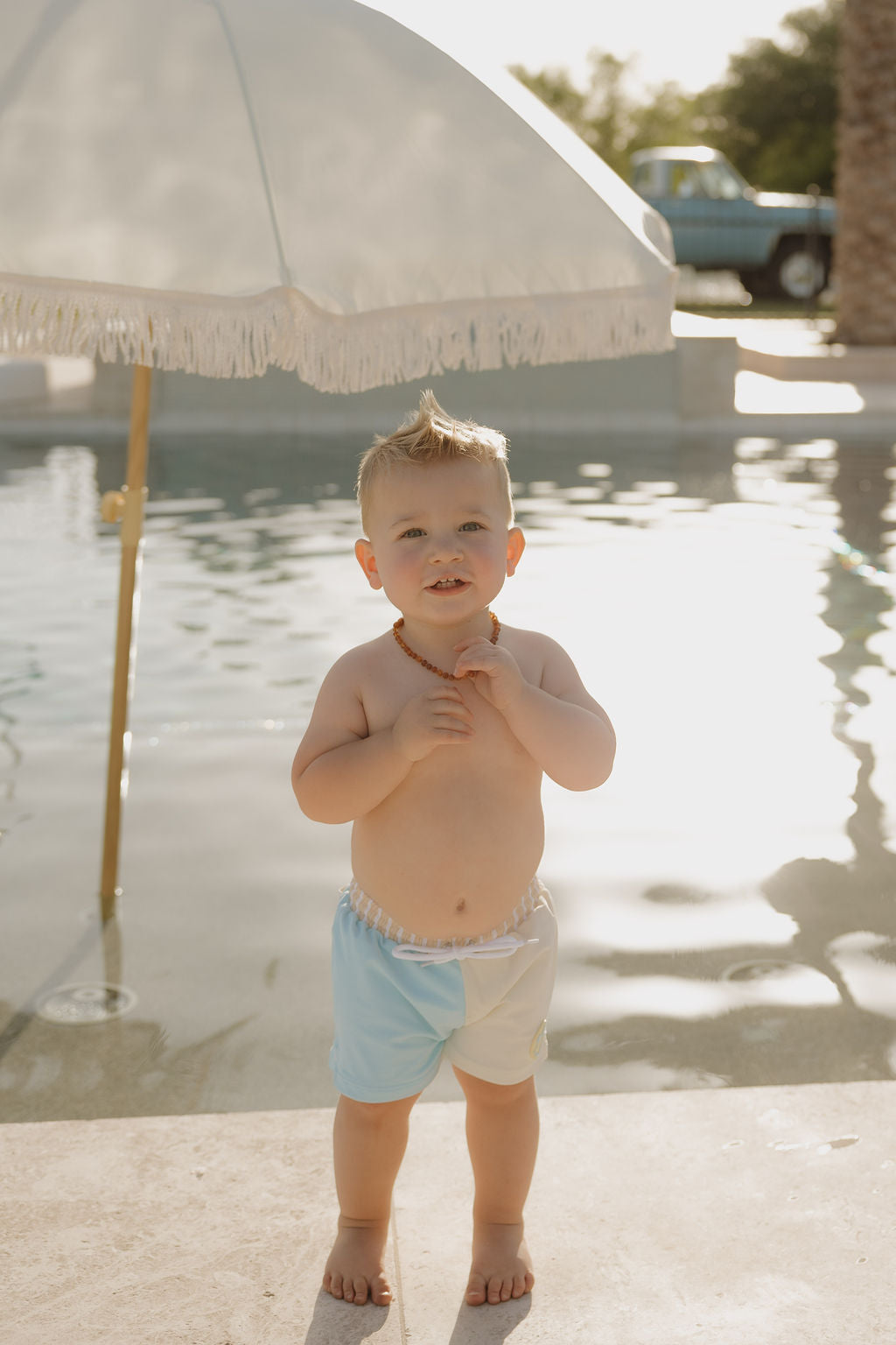 A toddler in lolo webb 2’s Child Board Short | Surf & Sand stands by a pool under a white fringed umbrella, smiling with hands at his chest. Sunlight sparkles on the water—part of our limited edition collection.