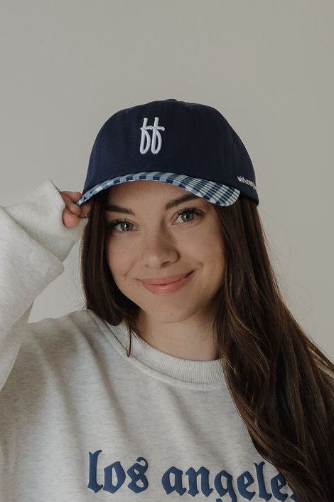 A young woman with long brown hair smiles, wearing a ff Blue baseball cap by forever french baby and a light grey “los angeles” sweatshirt. She poses against a plain background—spot clean with warm water to keep items fresh.