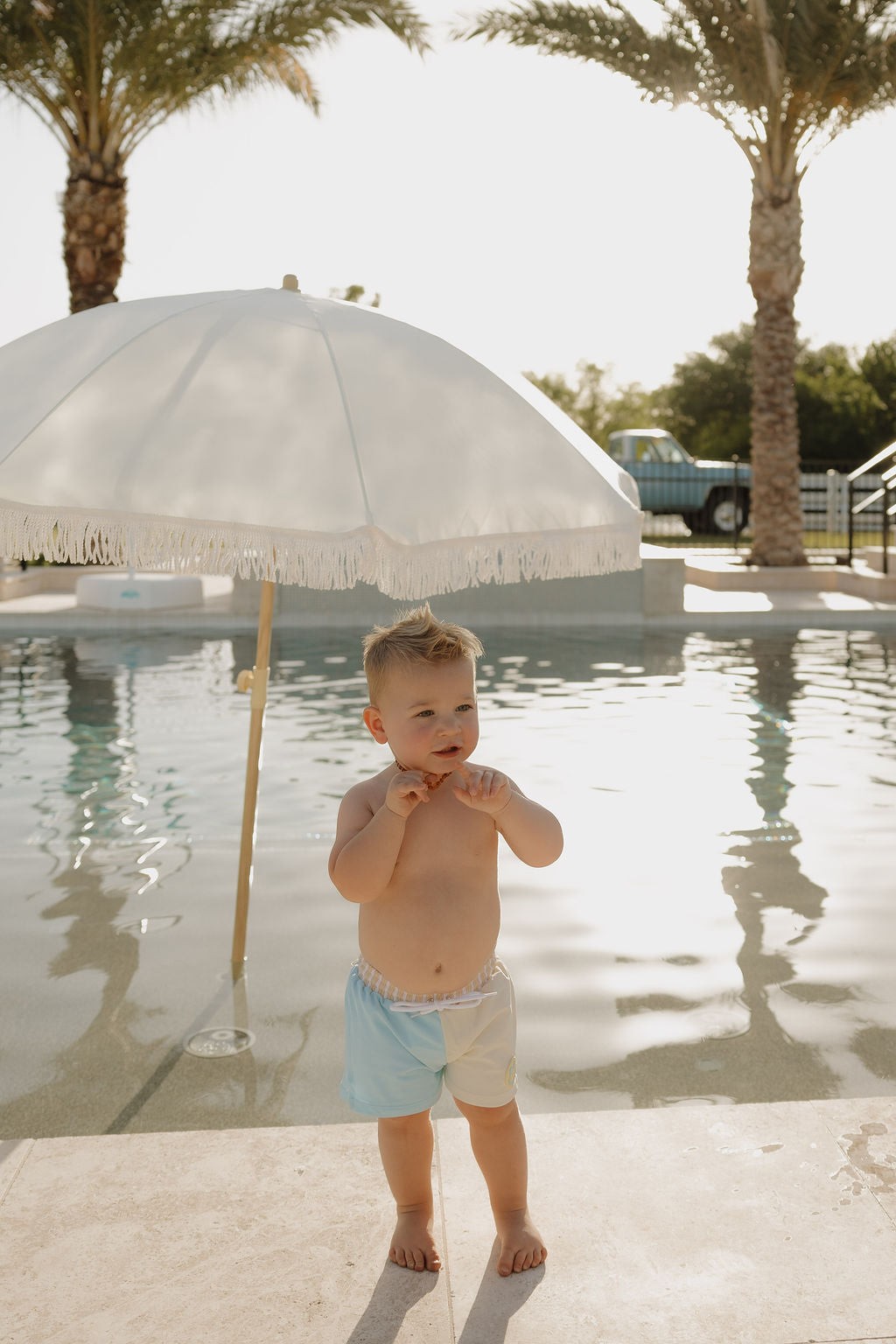 A child in lolo webb 2's limited edition Child Board Short | Surf & Sand stands poolside with a white fringed umbrella, palm trees, and a vintage truck in the background on a sunny day.