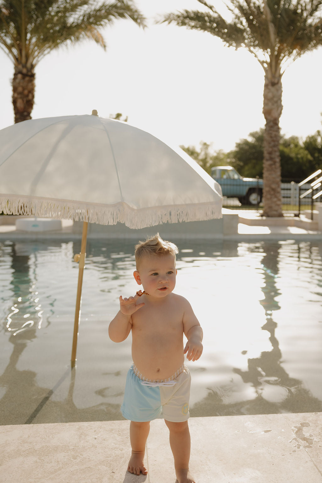 A toddler in lolo webb 2's Child Board Short | Surf & Sand stands poolside, waving under a white fringed umbrella with palm trees behind on a sunny day.