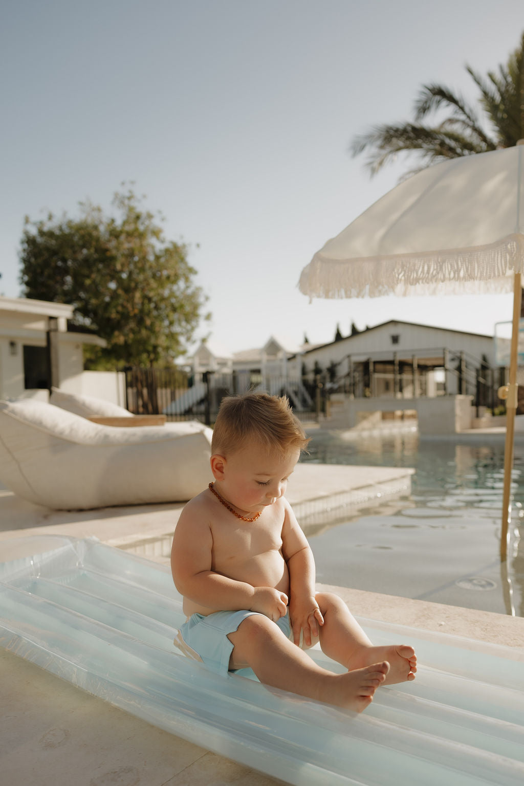 A young child wearing the lolo webb 2 Child Board Short | Surf & Sand sits on a blue pool float by the water, under a sunny sky with palm trees and white umbrellas—the perfect limited edition summer moment.