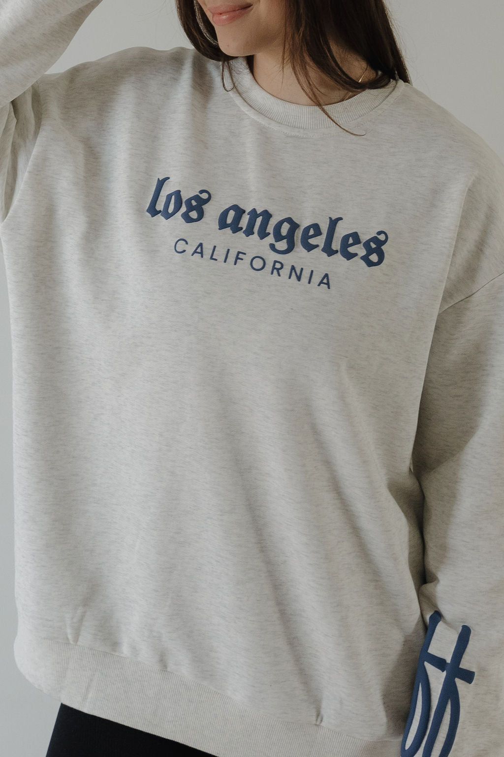 A woman models the forever french baby Adult Sweatshirt | ff Los Angeles in light gray with "Los Angeles California" in blue on the front. Only her lower face and upper body are visible.