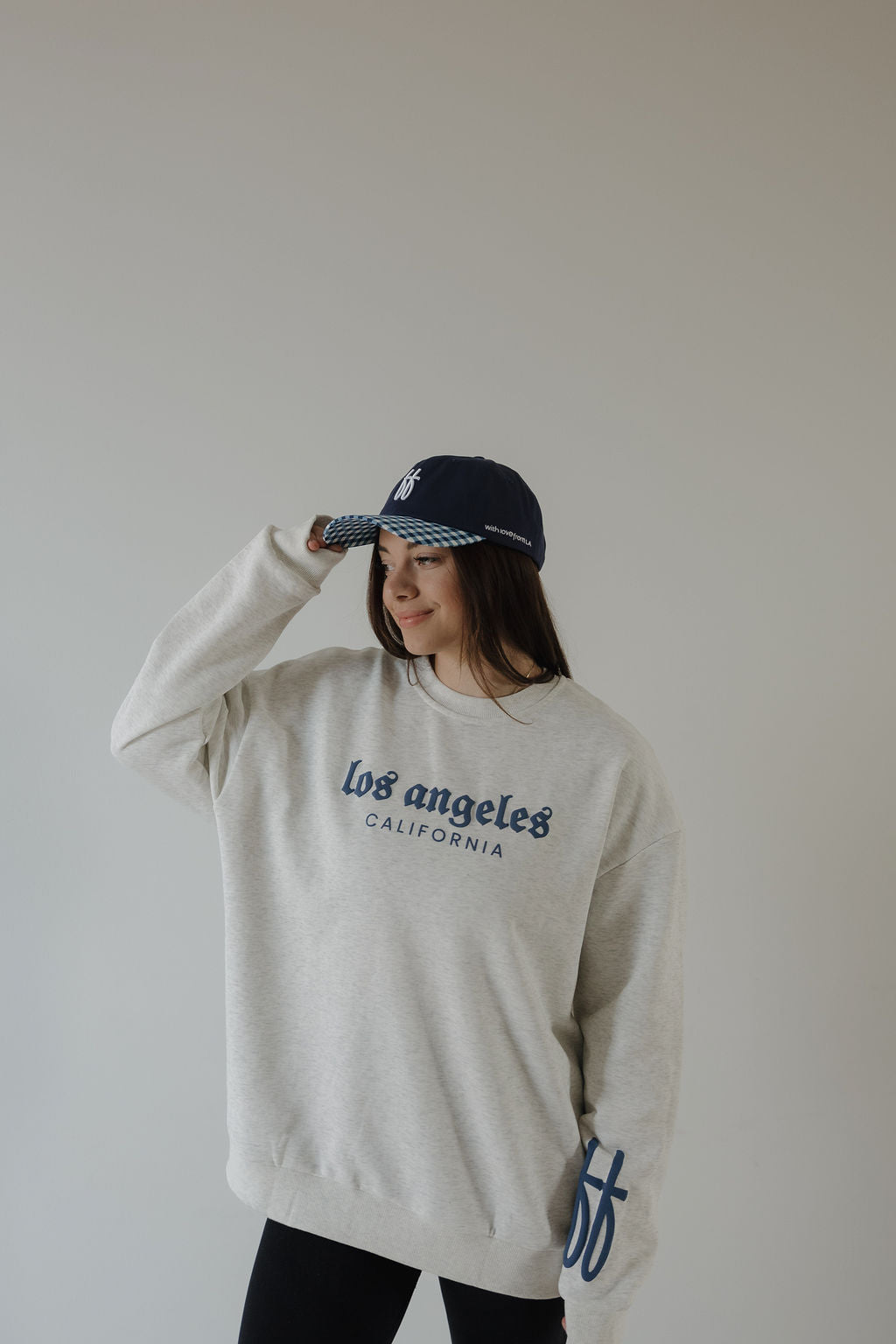 A person in a forever french baby ff Los Angeles adult sweatshirt and a navy blue cap stands against a plain light background, smiling and holding the brim of their cap.