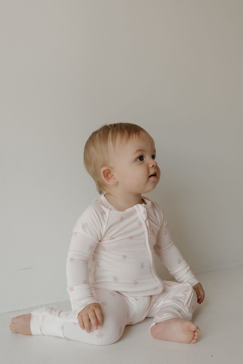A baby with light hair sits on the floor against a plain white wall, wearing the Bamboo Zip Pajamas | Rosie by forever french baby. This adorable outfit is crafted from hypoallergenic materials to ensure comfort and gentleness on sensitive skin.