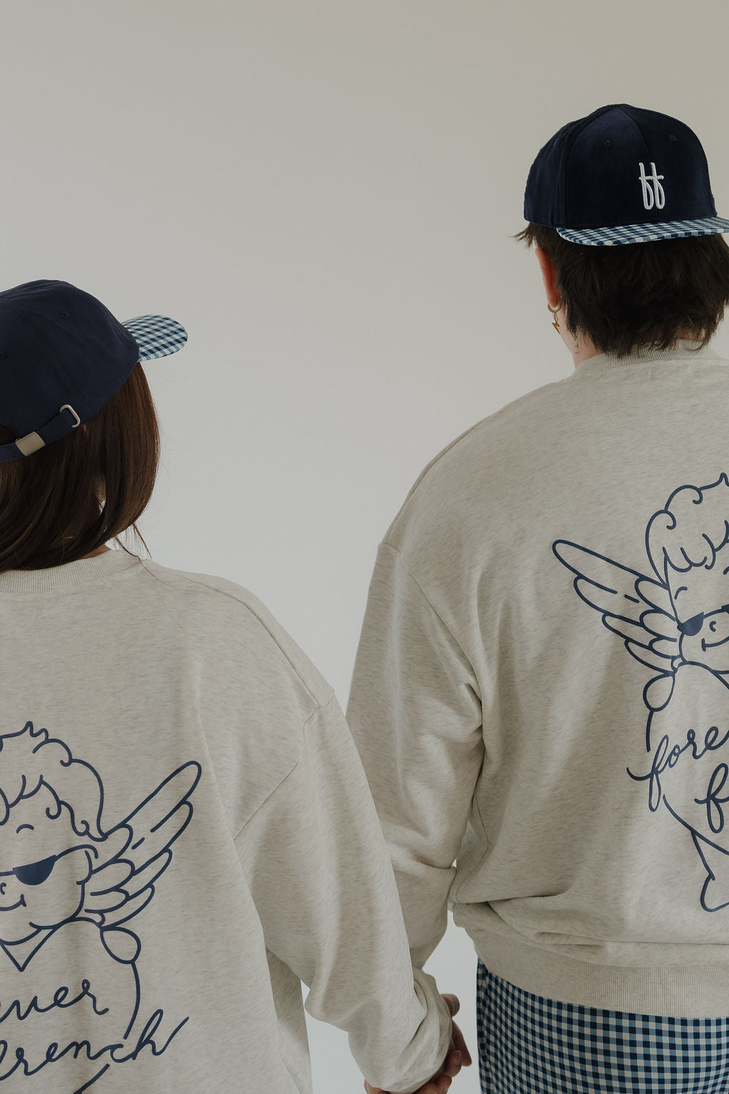 Two people in matching forever french baby Adult Sweatshirt | ff Los Angeles, featuring a blue cartoon angel and "forever french," with navy blue caps, hold hands and face away from the camera against a plain background.