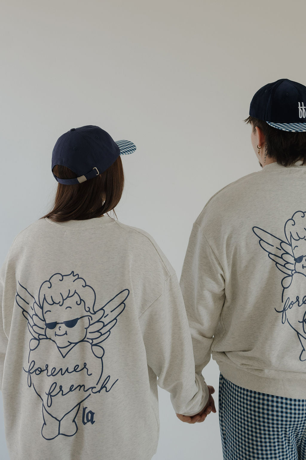 Two people are seen from behind, holding hands in matching forever french baby Adult Sweatshirt | ff Los Angeles—featuring a cartoon angel and "forever french" on the back—paired with blue caps, checkered bottoms, against a plain background.