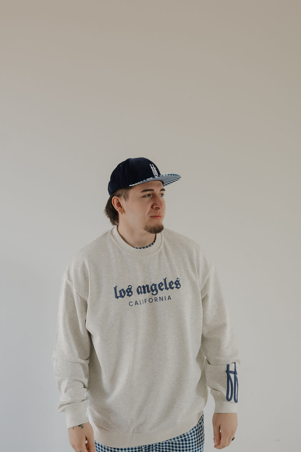 A man in a light gray forever french baby Adult Sweatshirt | ff Los Angeles, blue checkered pants, and a navy baseball cap stands against an off-white background, looking slightly to the side.