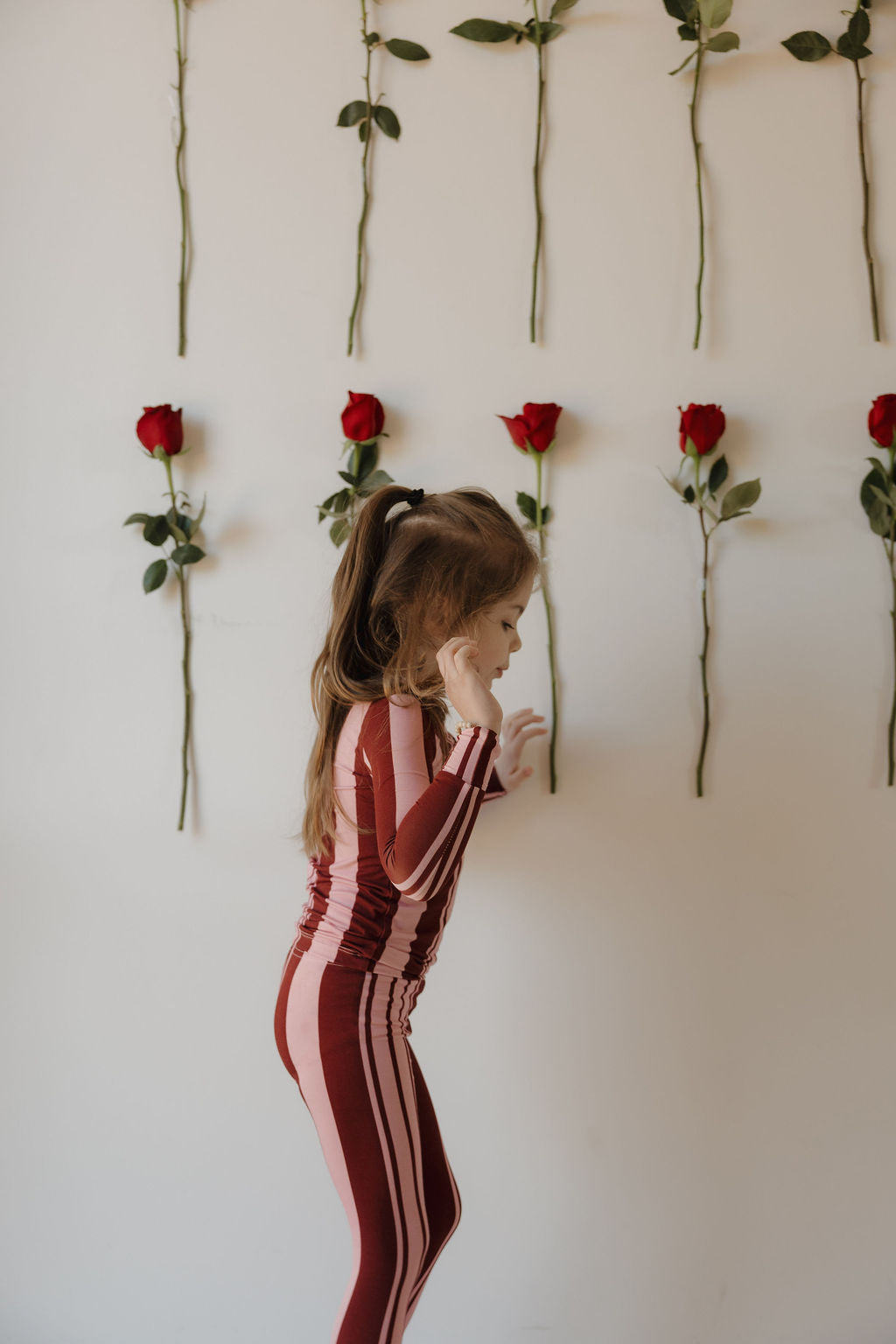 A young girl wears forever french baby's Bamboo Two Piece Pajamas | Sweetheart Stripes, standing sideways before a white wall adorned with vertical red roses.