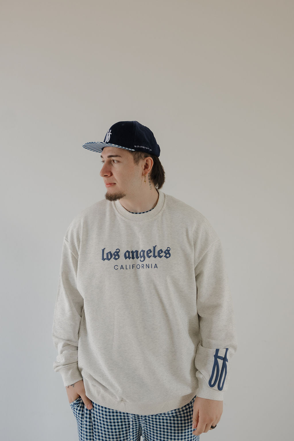 Someone wearing the forever french baby Adult Sweatshirt | ff Los Angeles in beige, blue plaid pants, and a navy baseball cap stands against a plain light background, looking to the side.