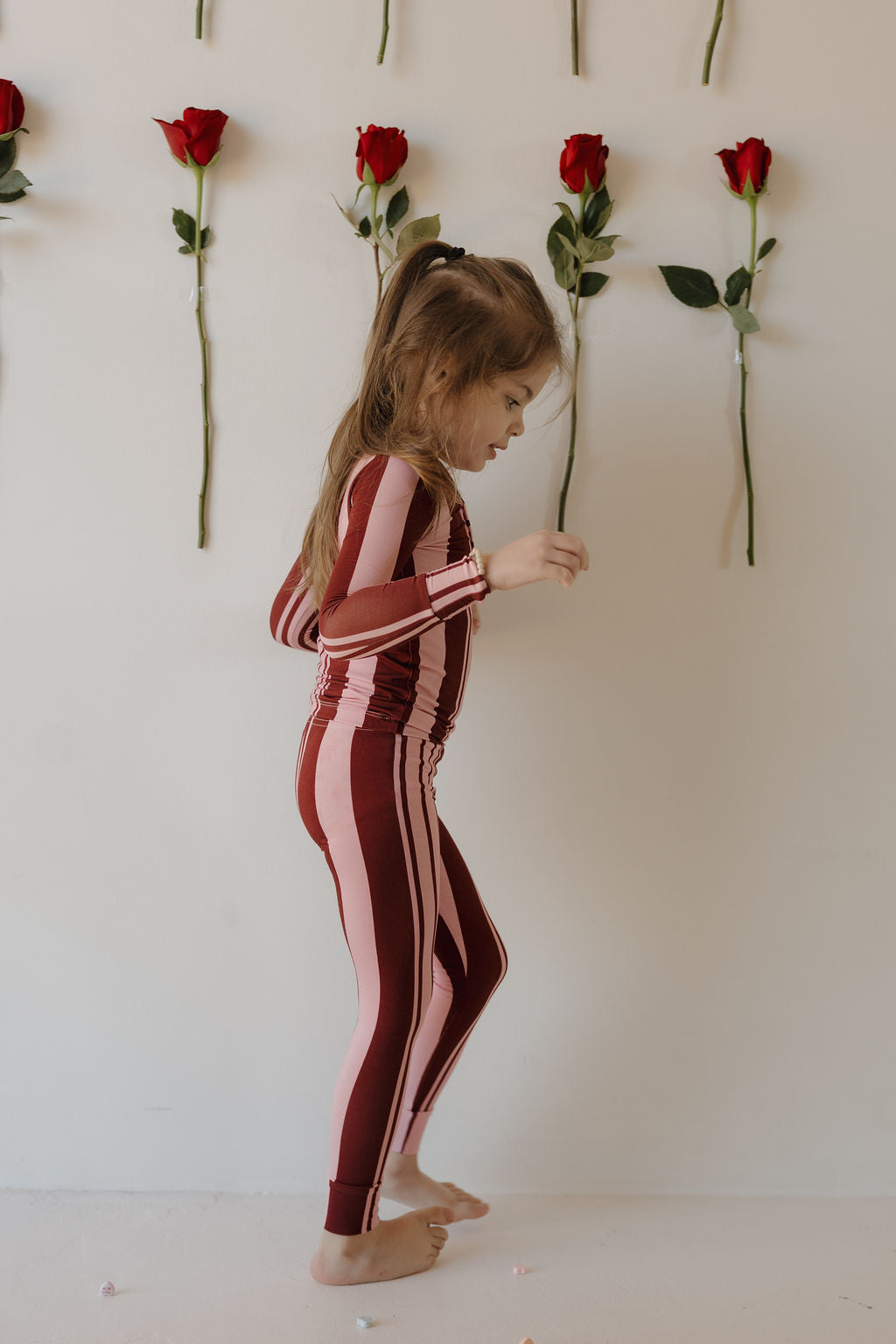 A young child wears forever french baby’s Bamboo Two Piece Pajamas in Sweetheart Stripes, standing sideways in front of a white wall adorned with vertical red roses.