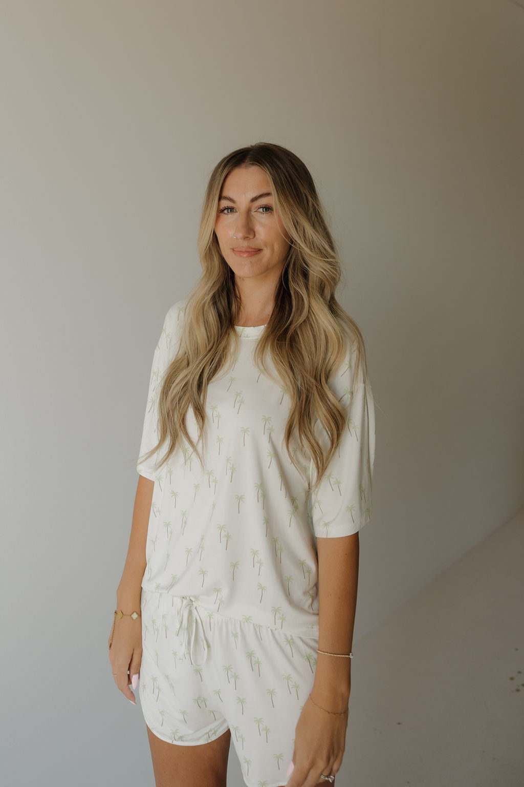 A woman with long, wavy blonde hair stands against a plain, neutral background wearing the forever french baby Women’s Bamboo Short Set | Palm—a breathable, hypo-allergenic pajama set featuring a subtle palm tree pattern and crafted from bamboo fabric.