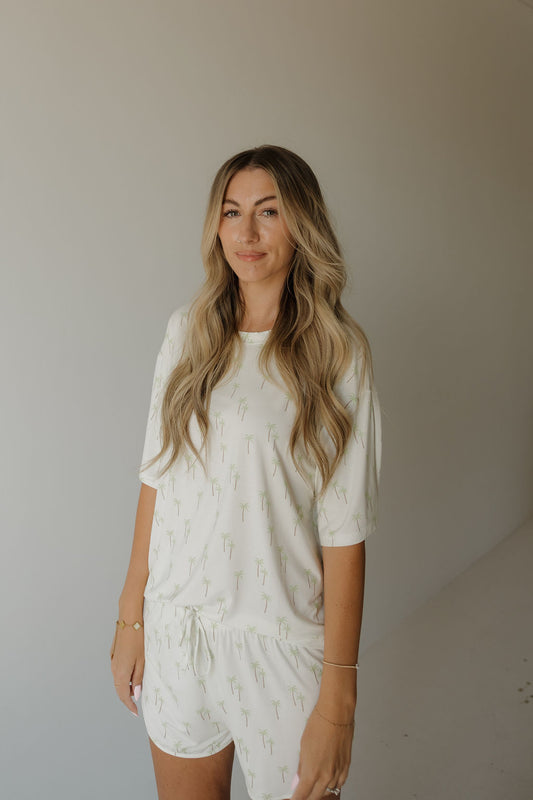 A woman with long, wavy blonde hair stands against a plain, neutral background wearing the forever french baby Women’s Bamboo Short Set | Palm—a breathable, hypo-allergenic pajama set featuring a subtle palm tree pattern and crafted from bamboo fabric.