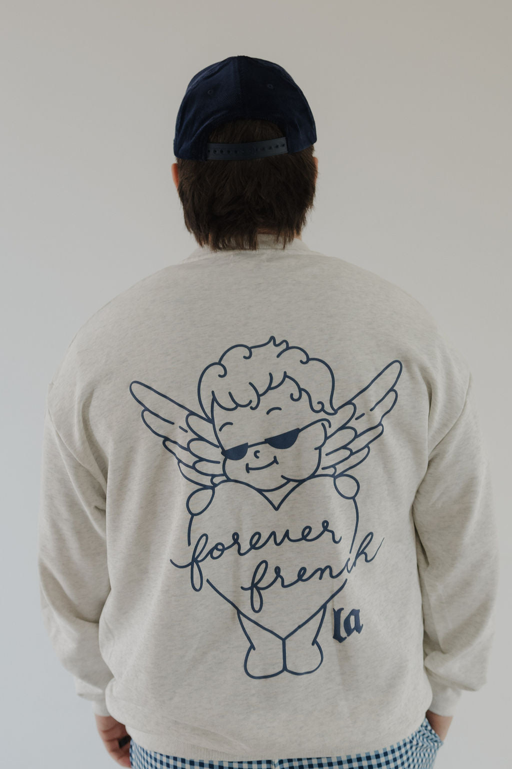 A person wearing the Adult Sweatshirt | ff Los Angeles by forever french baby, featuring a cartoon angel with a heart and "forever french" on the back. They are facing away, paired with a dark cap and checkered pants.