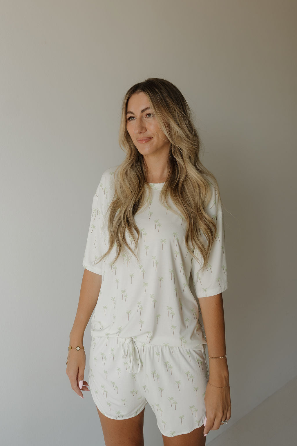 A woman with long wavy blonde hair wears the forever french baby Women’s Bamboo Short Set | Palm—a breathable bamboo pajama set featuring subtle small green palm tree prints—as she stands against a plain light-colored wall and smiles softly.
