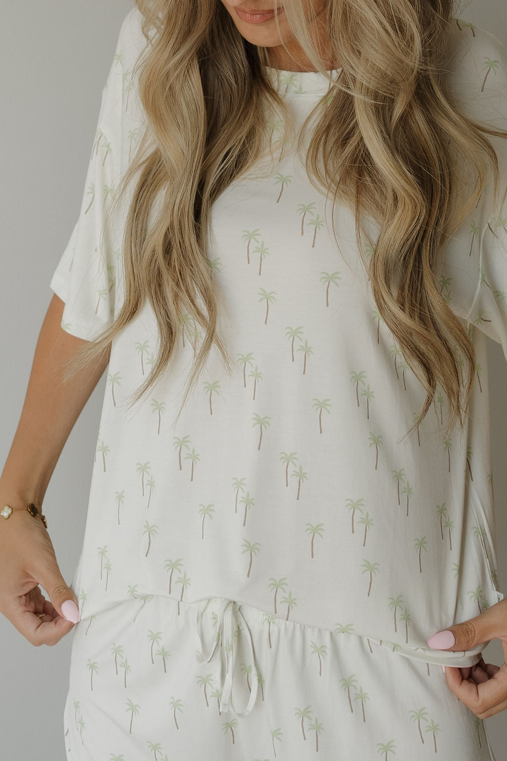 A woman with long blonde hair, wearing the forever french baby Women’s Bamboo Short Set in Palm—a matching white pajama set featuring light green palm tree prints and crafted from breathable bamboo—holds the hem of her shirt.