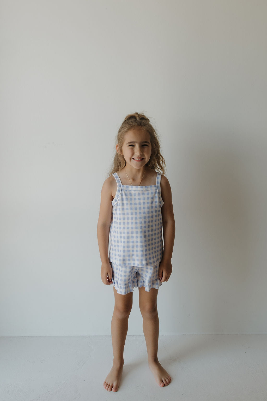 A young girl with long hair stands barefoot against a plain white wall, smiling and wearing the forever french baby Girl’s Ruffle Short Set | Marilyn, which features a charming blue and white gingham print tank and matching ruffle shorts.