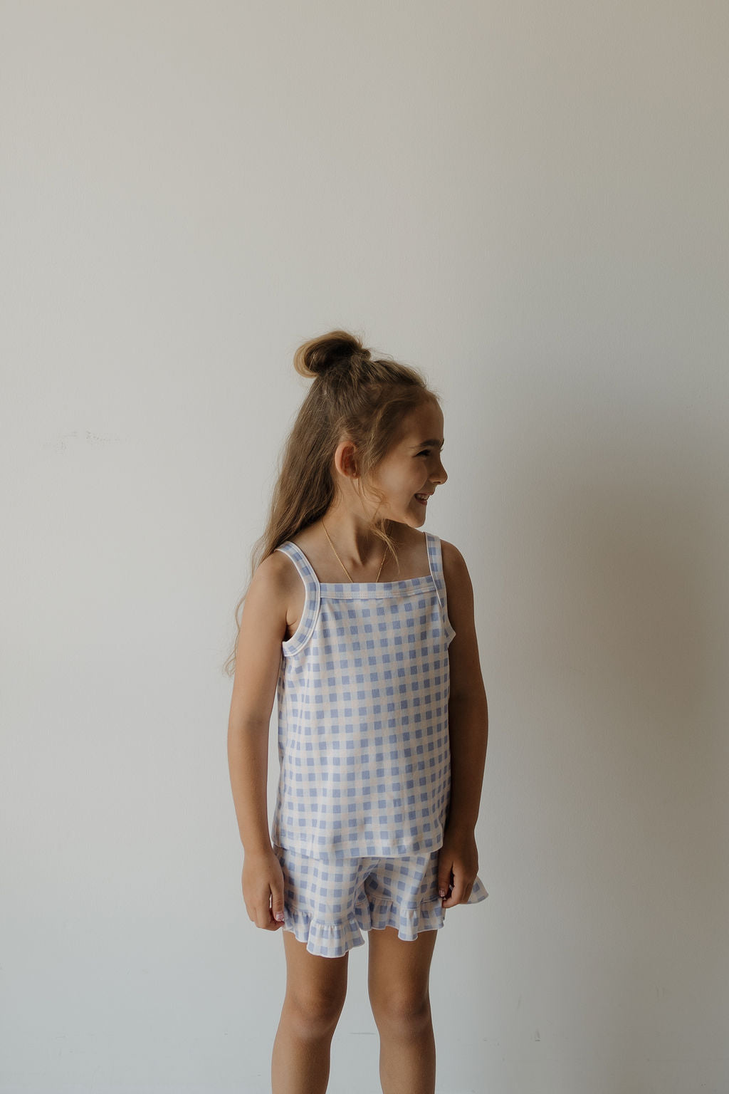 A young girl with long hair, wearing the Girl’s Ruffle Short Set | Marilyn by forever french baby, stands smiling to her left against a plain white background.