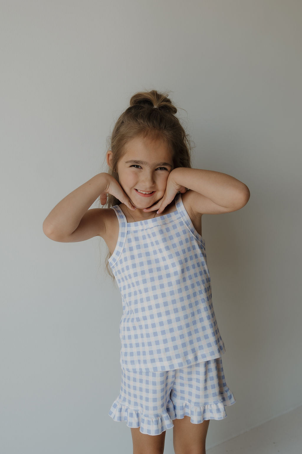 A young girl with long hair styled half-up poses playfully, smiling and touching her face. She wears the forever french baby Girl’s Ruffle Short Set | Marilyn in a blue and white gingham print cotton blend, standing against a plain, light-colored background.