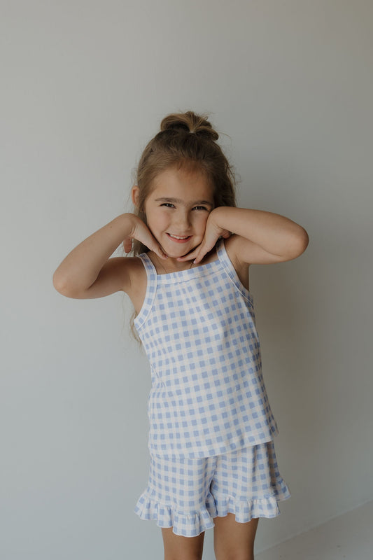 A young girl with long hair styled half-up poses playfully, smiling and touching her face. She wears the forever french baby Girl’s Ruffle Short Set | Marilyn in a blue and white gingham print cotton blend, standing against a plain, light-colored background.