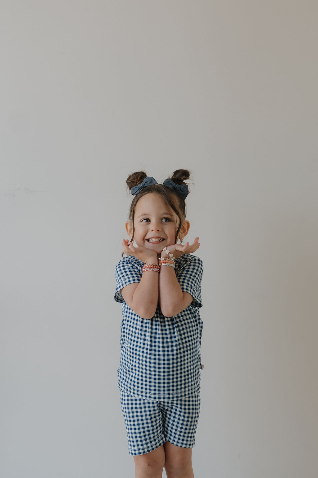 A smiling young child with brown hair in two buns wears the forever french baby Bamboo Short Set in Blue Gingham with a matching headband, posing against a plain light background.