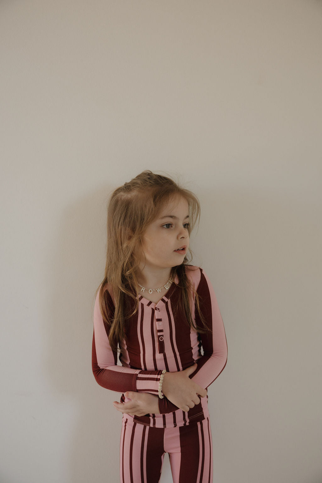 A young girl with long light brown hair stands against a plain wall, wearing forever french baby's Bamboo Two Piece Pajamas in Sweetheart Stripes—maroon and pink striped hypo-allergenic sleepwear—with arms folded, looking to the side.