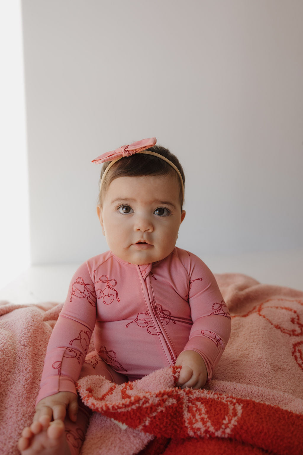 A baby wearing Minty Cherries Bamboo Zip Pajamas in Iconic Cherry with a matching headband sits on a pink and red blanket, gazing upward with a neutral expression.