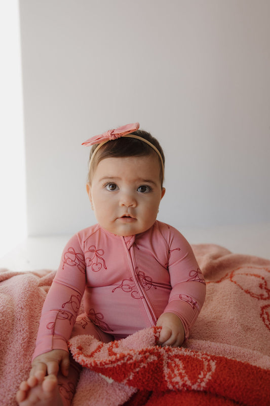 A baby wearing Minty Cherries Bamboo Zip Pajamas in Iconic Cherry with a matching headband sits on a pink and red blanket, gazing upward with a neutral expression.