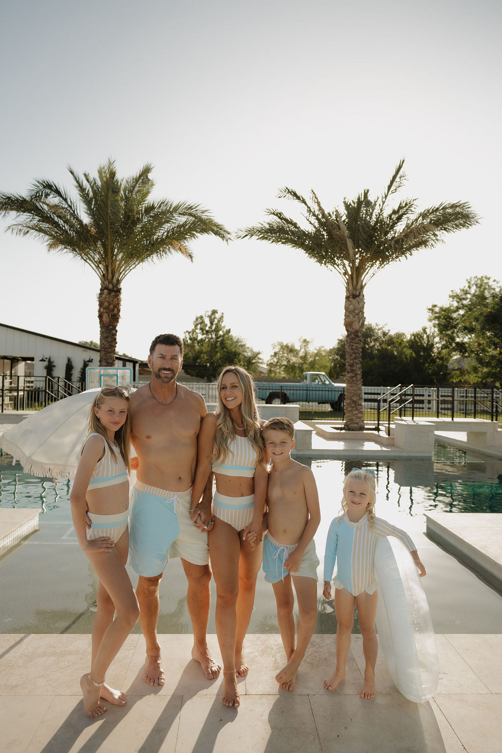 A family of five poses poolside in matching lolo webb 2 swimwear, with the kids wearing Child Board Short | Surf & Sand and the youngest holding a clear pool float among palm trees in the sun.