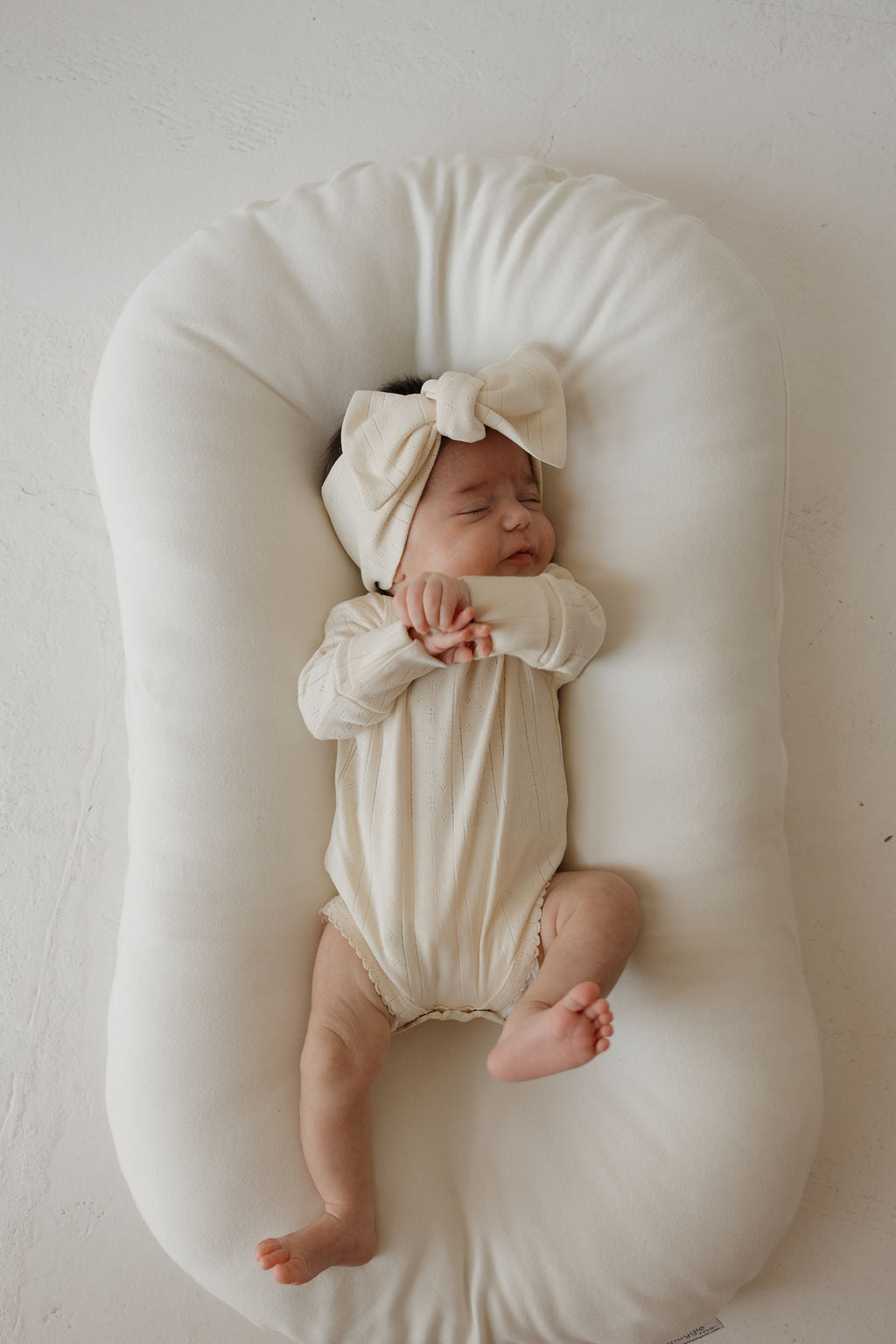 A baby wearing the Long Sleeve Snapsuit | Swan by forever french baby and a large matching headband sleeps peacefully on a soft, white cushioned pad.