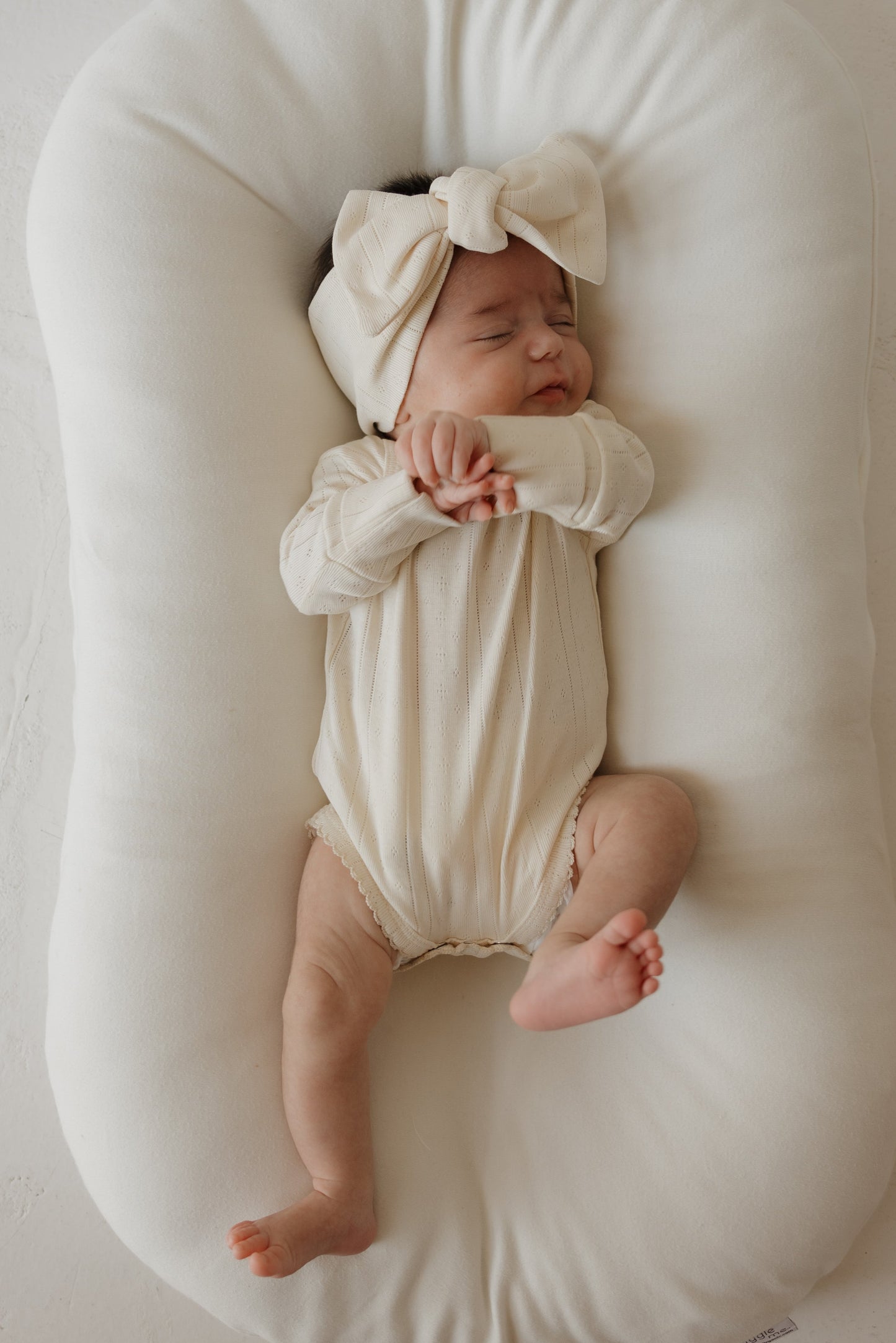 A baby wearing the forever french baby Long Sleeve Snapsuit | Swan and a matching bow headband sleeps peacefully on a soft, white cushioned pad.
