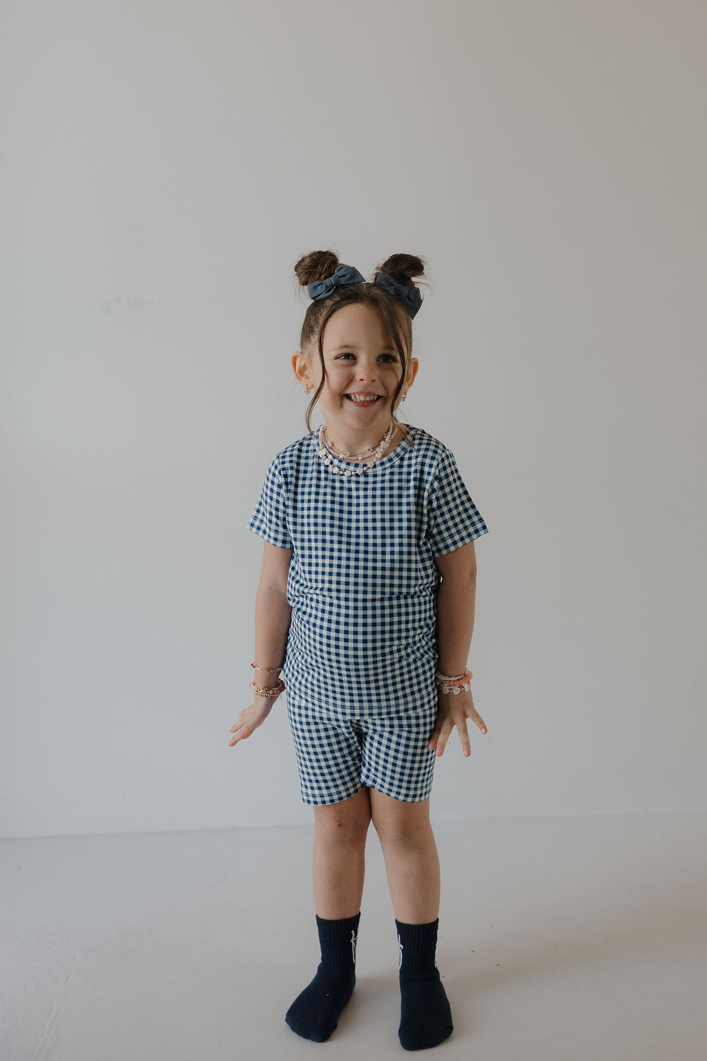 A smiling young child with two buns wears the forever french baby Bamboo Short Set in Blue Gingham, paired with navy socks and beaded bracelets, standing against a plain light background.