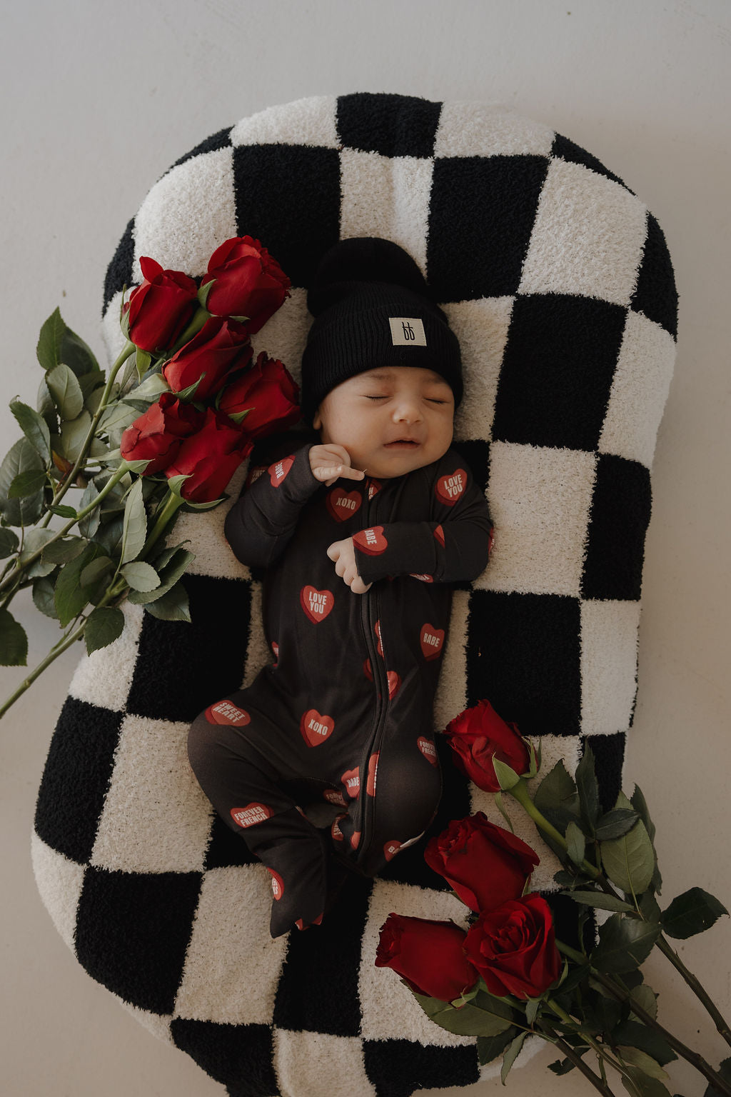 A baby in forever french baby's Bamboo Zip Pajamas | Love Notes – Charcoal—a hypoallergenic black onesie with red heart patterns and a black beanie—lies on a checkered cushion, surrounded by red roses.