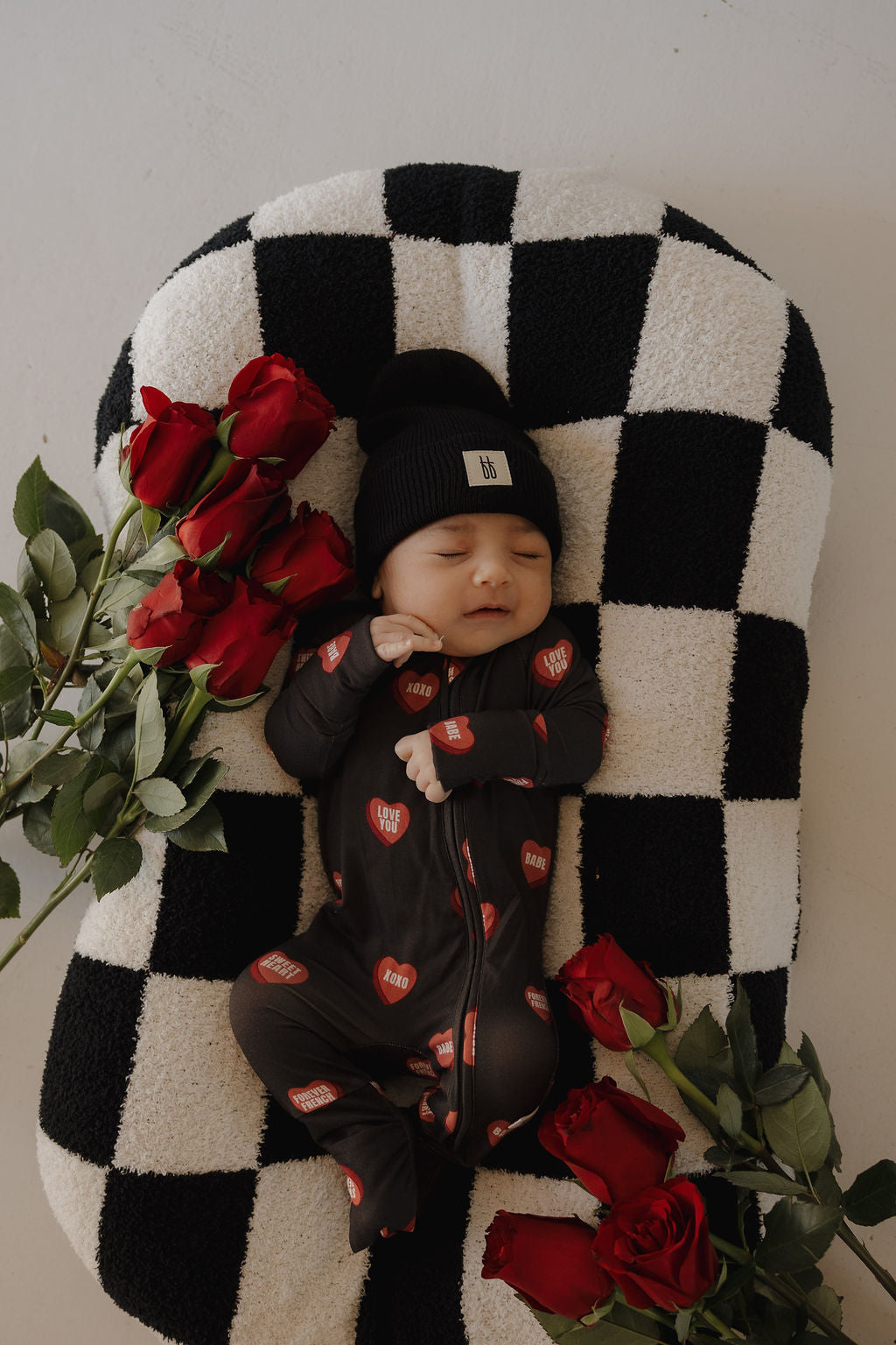 A baby in forever french baby's Bamboo Zip Pajamas | Love Notes – Charcoal and a black beanie sleeps on a black-and-white checkered cushion, surrounded by red roses.