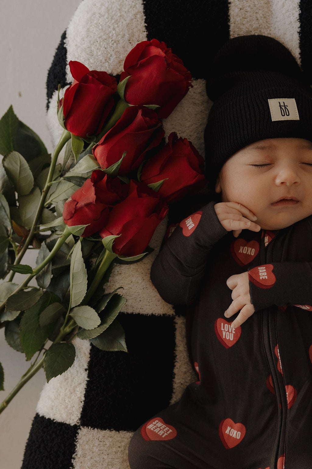 A baby in forever french baby Bamboo Zip Pajamas | Love Notes – Charcoal and a black beanie lies on a black-and-white striped blanket beside red roses.