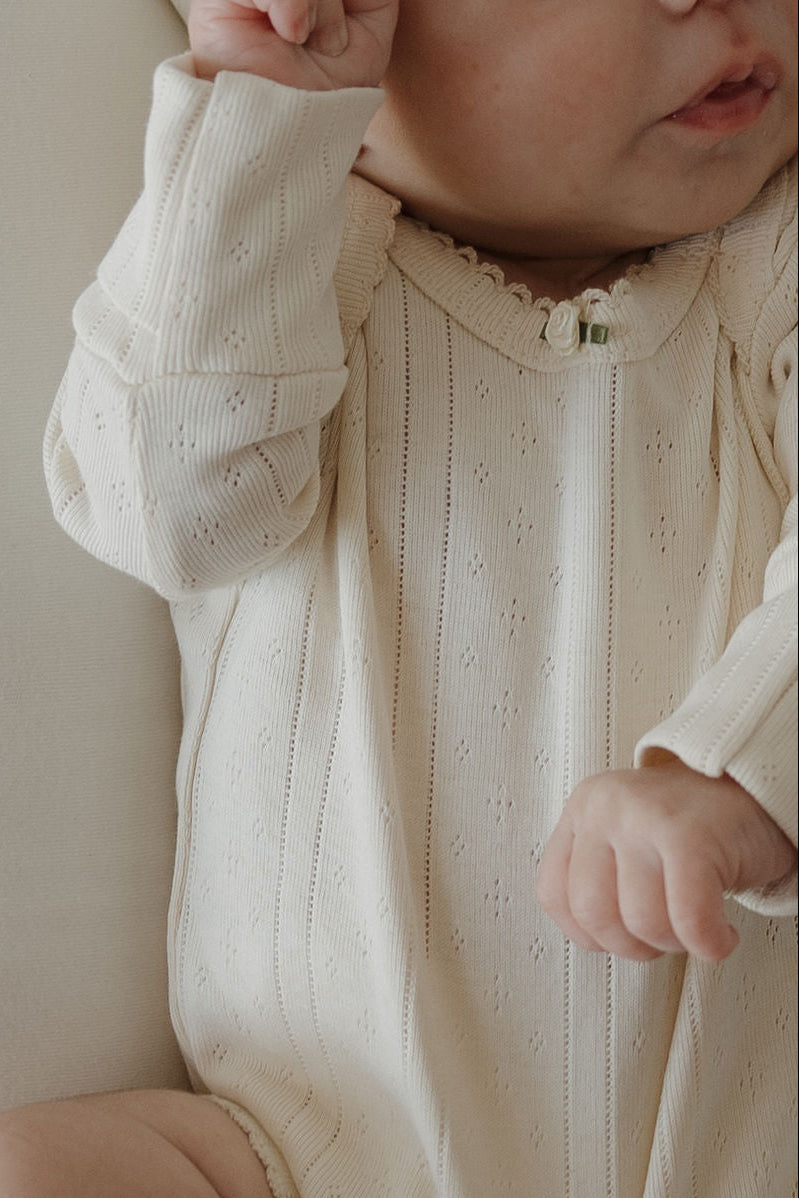 A baby wearing the forever french baby Long Sleeve Snapsuit | Swan—featuring cream-colored, textured pointelle cotton and long sleeves—sits with only the lower half of their face and body visible, one arm raised and the other bent.