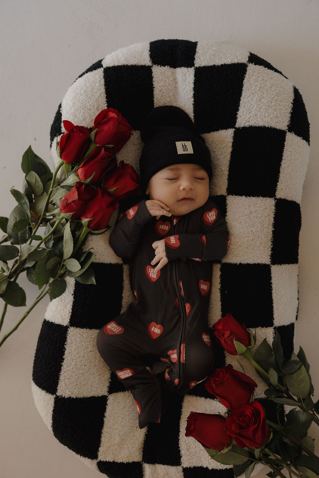 A baby in forever french baby’s Bamboo Zip Pajamas | Love Notes – Charcoal and a black hat sleeps on a black-and-white checkered pillow, surrounded by red roses.