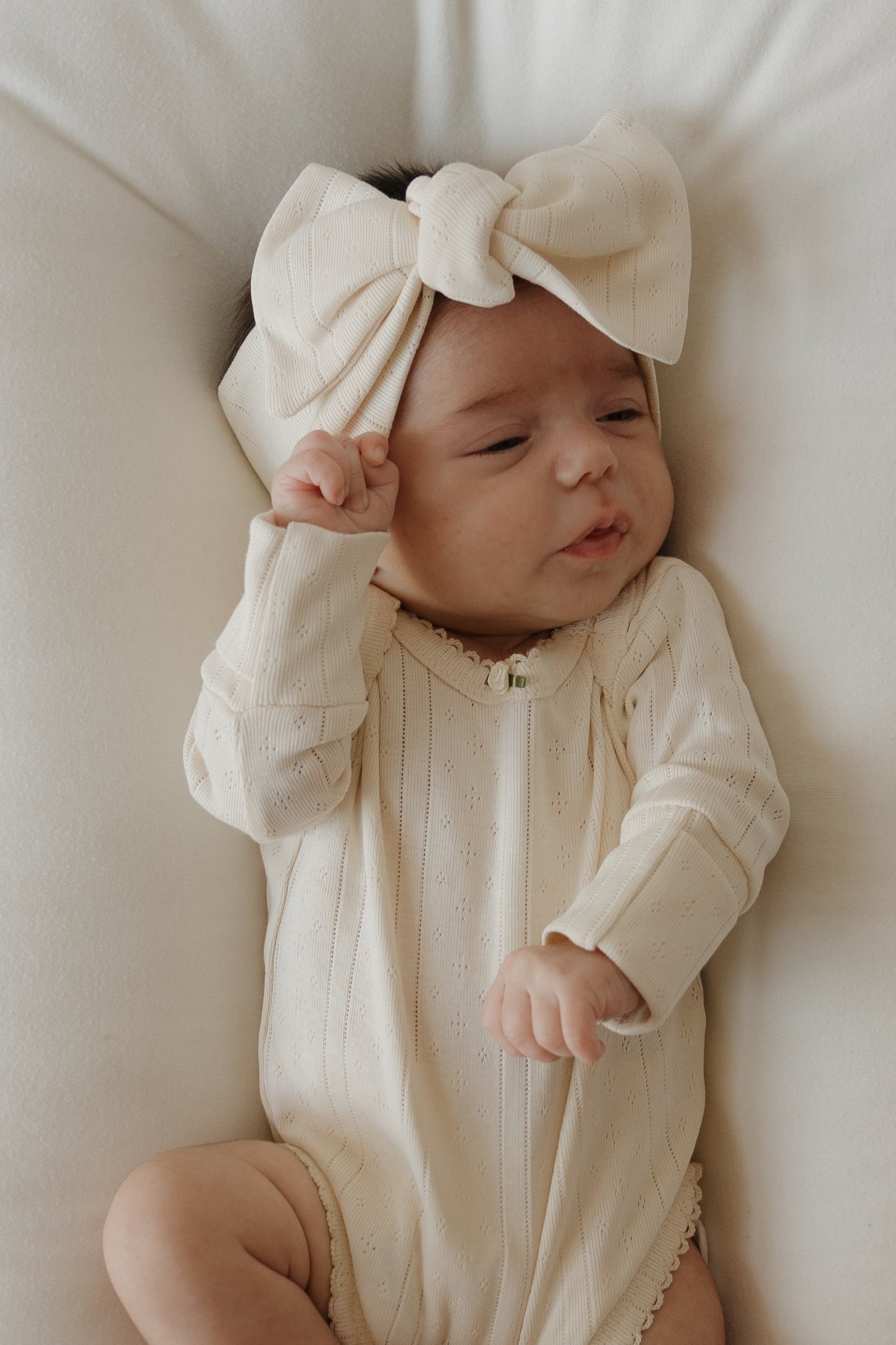 A baby wearing a cream-colored pointelle cotton outfit and the forever french baby Pointelle Head Wrap | Swan lies on a white blanket, raising one hand near their head and making a playful facial expression.