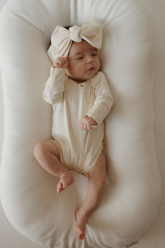 A baby wearing the forever french baby Long Sleeve Snapsuit | Swan, a cream-colored pointelle cotton outfit, and a large matching bow headband lies on a soft, white cushioned pad with one arm raised and eyes partially closed.