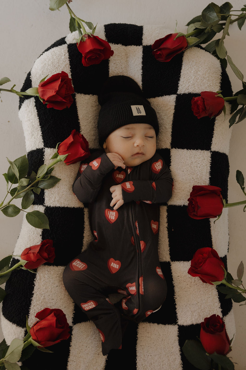 A baby sleeps peacefully in forever french baby's Bamboo Zip Pajamas | Love Notes – Charcoal, featuring red "LOVE" patches, with a matching black beanie on a checkered blanket surrounded by red roses.