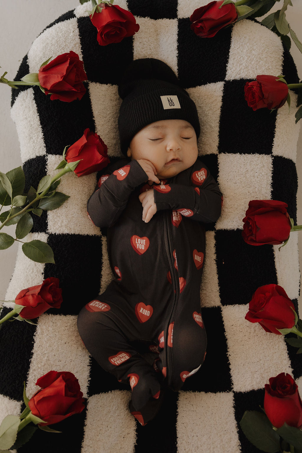 A baby in forever french baby’s Bamboo Zip Pajamas | Love Notes – Charcoal and a black beanie sleeps on a black-and-white checkered blanket, surrounded by red roses.