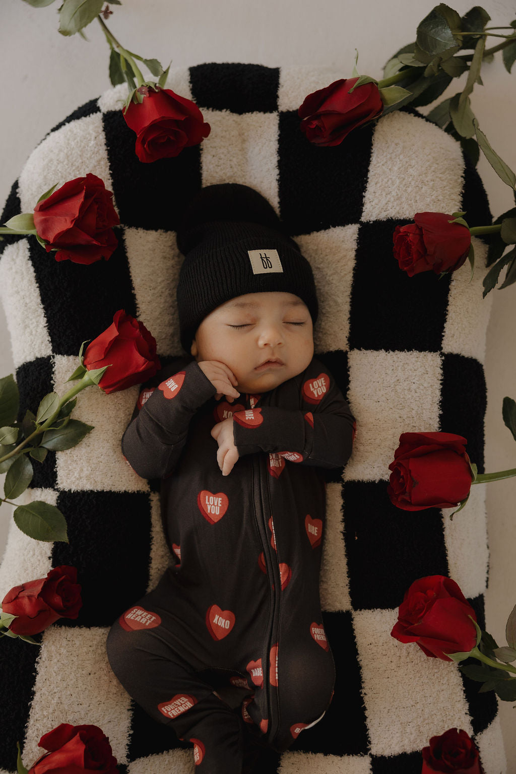 A baby in forever french baby Bamboo Zip Pajamas | Love Notes – Charcoal and a black beanie lies on a black-and-white checkered blanket with red "Love You" hearts, surrounded by red roses.